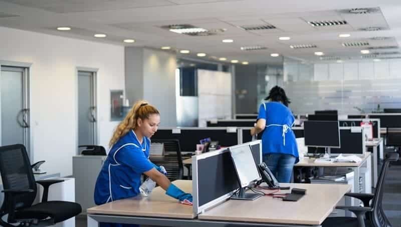 A woman in blue shirt cleaning desk near two other women.